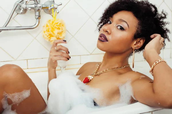 Young afro-american teen girl laying in bath with foam, wearing swag jewelry flawless, making selfie, modern lifestyle concept, rich woman