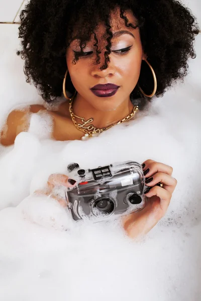 Young afro-american teen girl laying in bath with foam, wearing swag jewelry flawless, making selfie