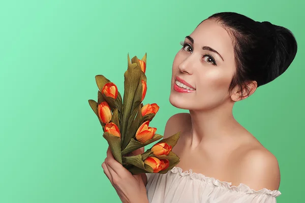 Spring beauty model studio shooting. Portrait of smiling young woman brunette  with flowers orange tulips on green background. Fashion fresh makeup. Perfect skin. Tenderness. Romantic style.