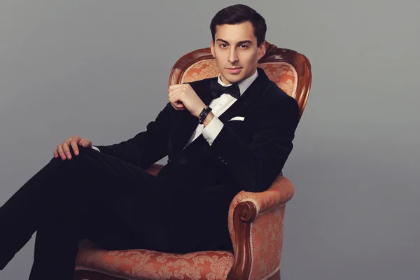 Confident successful young handsome man businessman in elegant suit with bow tie sitting on vintage armchair on grey background. Manhood. Male beauty. Fashion model studio shot. Italian style. Luxury.