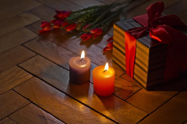 Burning candles, open gift box present with red bow and beige brown strips, bouquet of red scarlet flowers tulips on wooden background. Romantic style. Holidays.