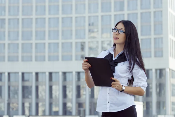 Young smiling beautiful elegant business woman in white blouse and black glasses with folder on background skyscraper.