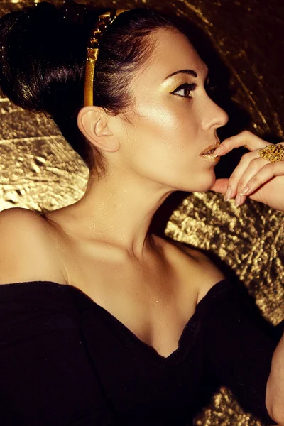 Young beautiful woman brunette with retro hairstyle, hoop looking into the distance. Gold background. Model shooting. Girl with gold fashion make-up. Luxury. Elegance. Sexuality. Sensuality.