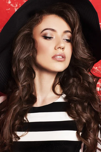 Portrait of beautiful young woman with long curly hair in black hat and striped dress on red background.