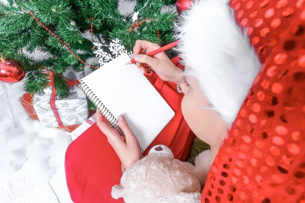 Girl in Santa hat writes letter to Santa near Christmas tree.