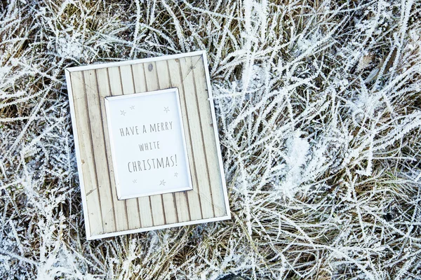 Old wooden frame over frozen grass. Have a merry white christmas