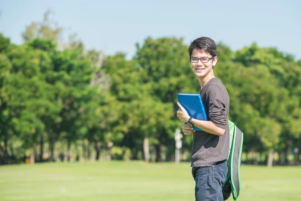 Asian student holding books and smiling while standing in park a