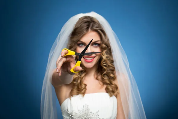 Young beautiful girl in wedding dress veil with scissors in hands on blue background