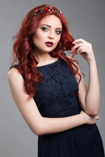 Beautiful red-haired fashion model posing in evening dress and in the diadem over dark background. female gestures of seduction. body language. touching her hair.