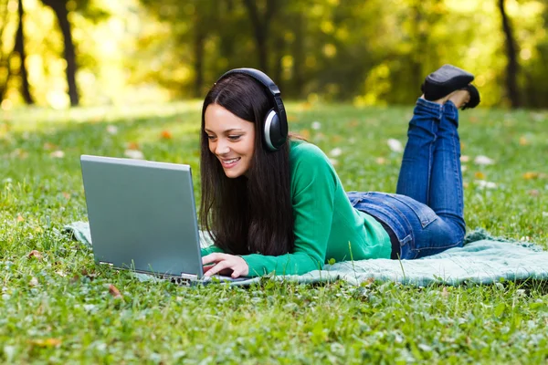 Woman with headphones is using her laptop while lying down in the nature