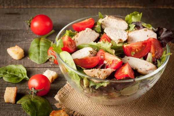 Fresh salad made of tomato, ruccola, chicken breast, arugula, crackers and spices. Caesar salad in a white, transparent bowl on wooden background