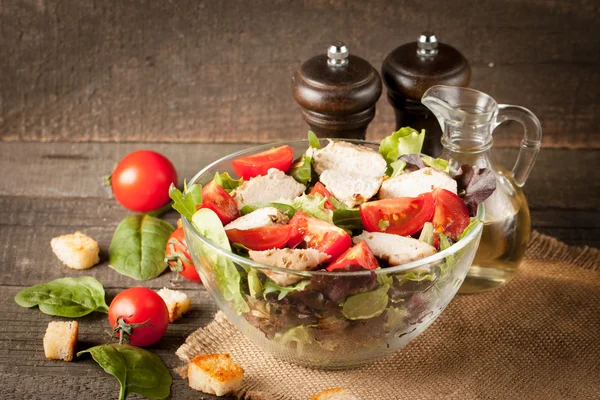 Fresh salad made of tomato, ruccola, chicken breast, arugula, crackers and spices. Caesar salad in a white, transparent bowl on wooden background