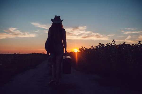 Girl in a cowboy hat standing with a suitcase on the road in the sunflower field. Waiting for help. Sunset.