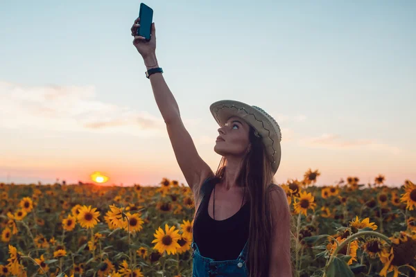 Girl in a cowboy hat lost in sunflower field catches a cellular phone. Sunset.