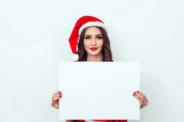 Beautiful girl in the hat of Santa Claus. Hold a sheet of paper. Red manicure. Red lips.