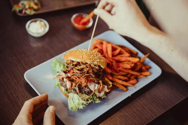 Man holds burger with hands and sweet potato fries  and dips on