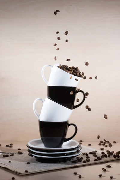 Roasted coffee beans falling down into black and white cups on the stack of the plates standing on newspaper.