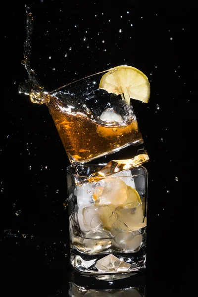 Falling piece of ice in one high quality glass of whiskey with many splashes standing on another glass full of ice and lemon slice.