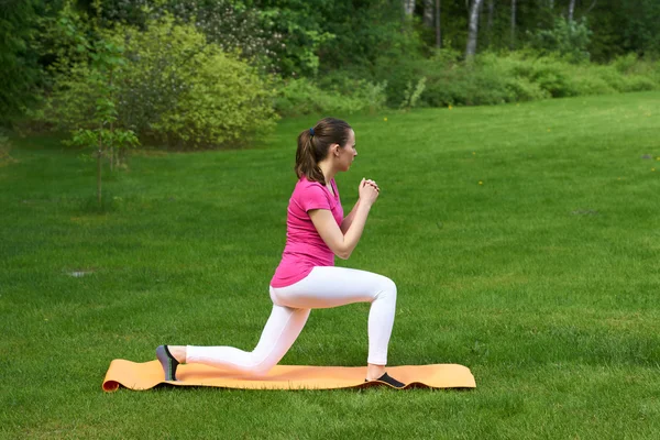 Sexy brown haired woman doing physical exercises for buttocks and legs on orange mat in outdoor on grass in nature.