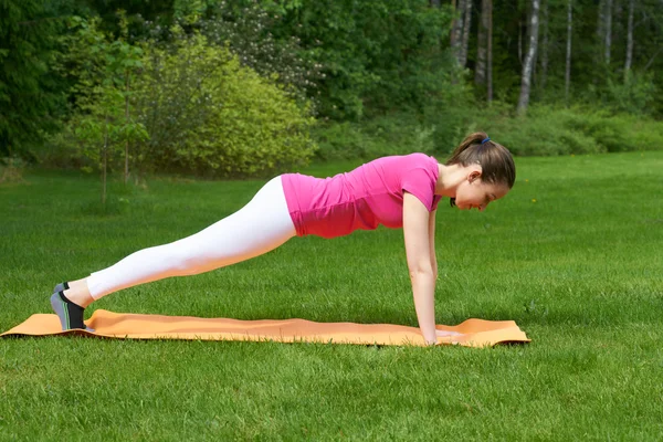 Beautiful brown haired woman doing exercises for muscles of hands, legs and stomach in plank pose on orange mat in outdoor grass in nature.