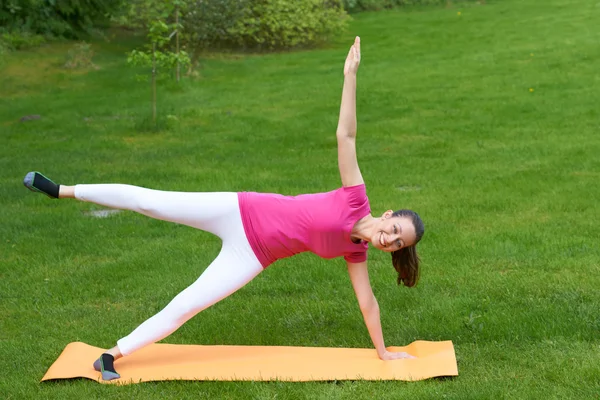 Happy brown haired woman pulling up one hand and leg and standing on others hand and leg on orange mat in outdoor grass in nature.