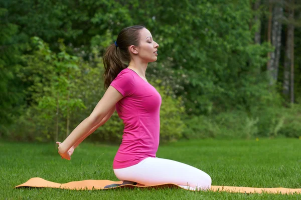 Beautiful brown haired woman with closed eyes doing stretching with hands stretched to backside on orange mat in outdoor in nature.