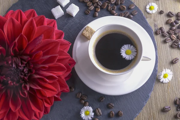 Composition made from cup of black tea with a black and brown background. Cup of black coffee with red flower, sugar, chamomile and coffee beans. It simbolizes nice morning with tasty tea.