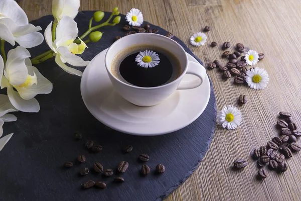 Composition made from cup of black tea with a black and brown background. Cup of black coffee with white flower, chamomile and coffee beans. It simbolizes nice morning with tasty tea.