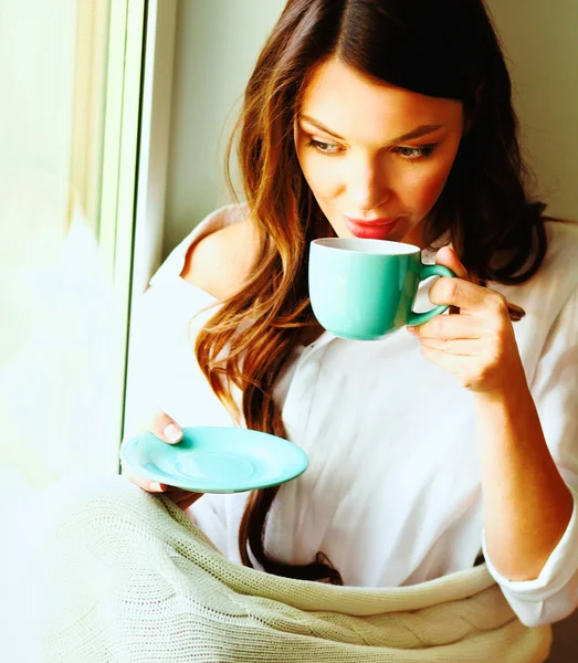 Young woman at home sitting near window relaxing in her living room reading book and drinking coffee or tea