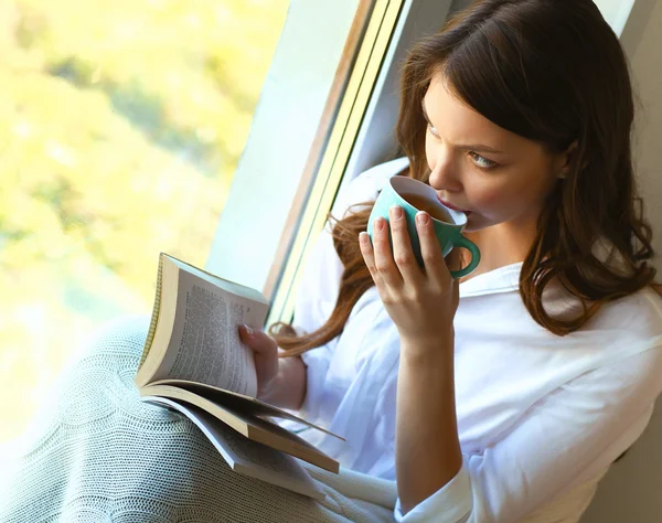 Young woman at home sitting near window relaxing in her living room reading book and drinking coffee or tea
