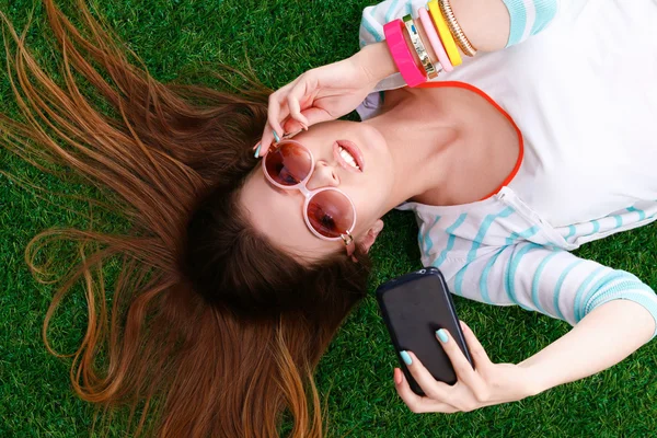 Beautiful young woman making selfie by her phone while lying in green grass