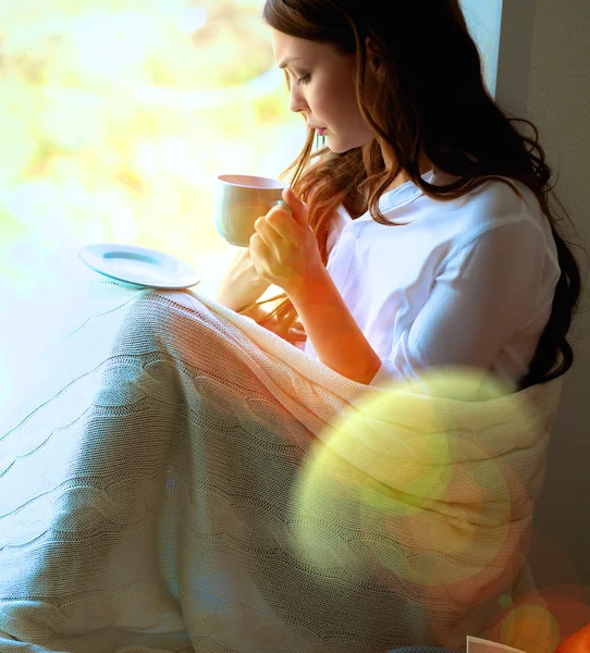 Young woman at home sitting near window relaxing in her living room reading book and drinking coffee or tea