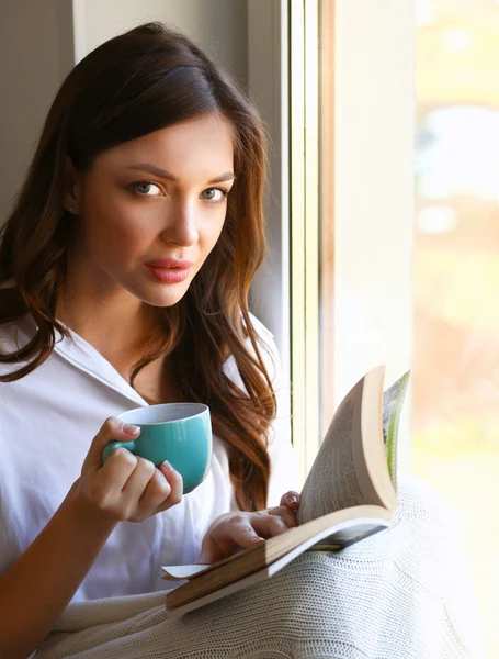 Young woman at home sitting near window relaxing in her living room reading book and drinking coffee or tea