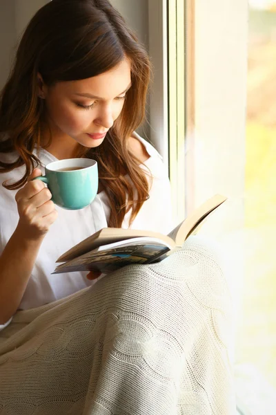 Young woman at home sitting near window relaxing in her living room reading book and drinking coffee or tea