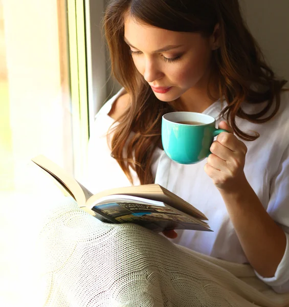 Young woman at home sitting near window relaxing in her living room reading book and drinking coffee or tea