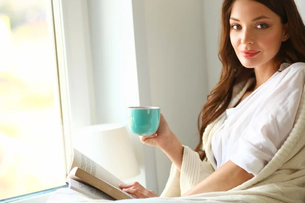 Young woman at home sitting near window relaxing in her living room reading book and drinking coffee or tea