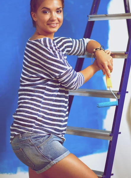 Happy beautiful young woman doing wall painting, standing near ladder