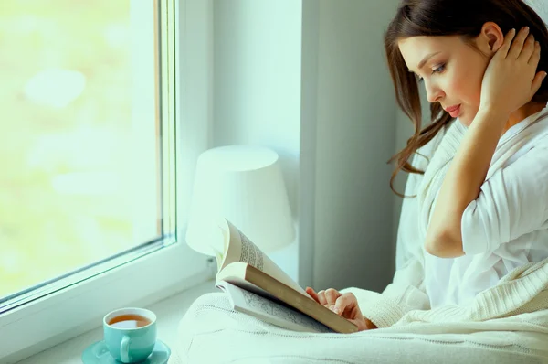 Young woman at home sitting near window relaxing in her living room reading book and drinking coffee or tea