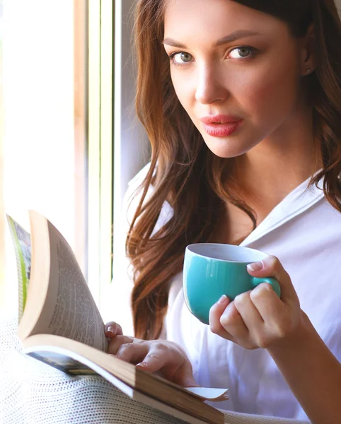 Young woman at home sitting near window relaxing in her living room reading book and drinking coffee or tea