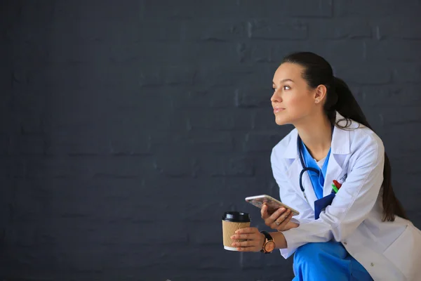 Female doctor sitting with mobile phone and drinking coffee