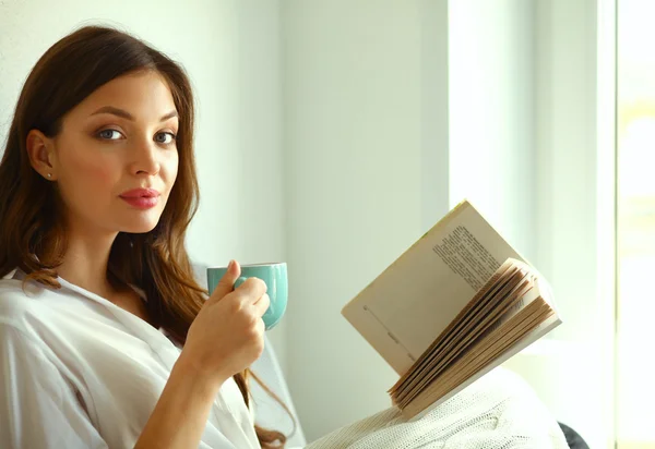 Young woman at home sitting near window relaxing in her living room reading book and drinking coffee or tea