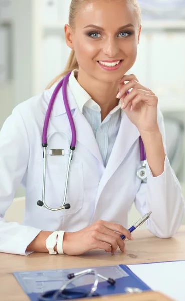 Beautiful young smiling female doctor sitting at the desk and writing.