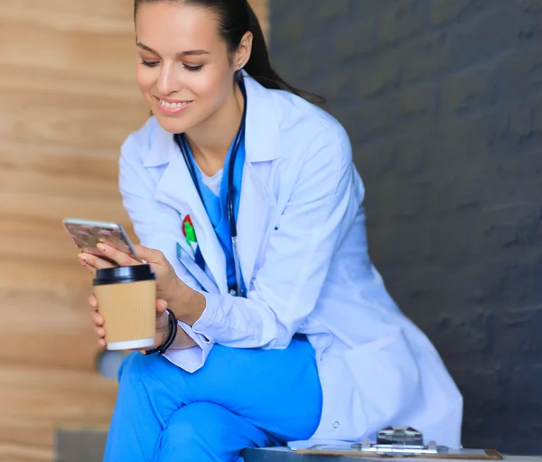 Female doctor sitting with mobile phone and drinking coffee