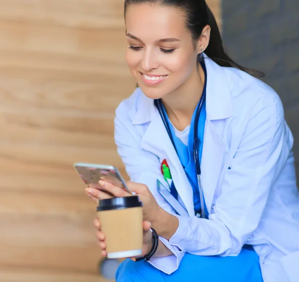 Female doctor sitting with mobile phone and drinking coffee