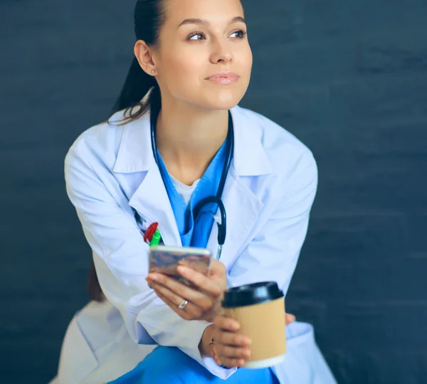 Female doctor sitting with mobile phone and drinking coffee