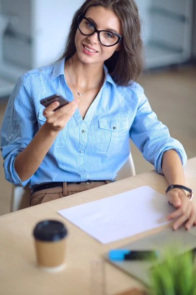 Beautiful young business woman sitting at office desk and holding cell phone