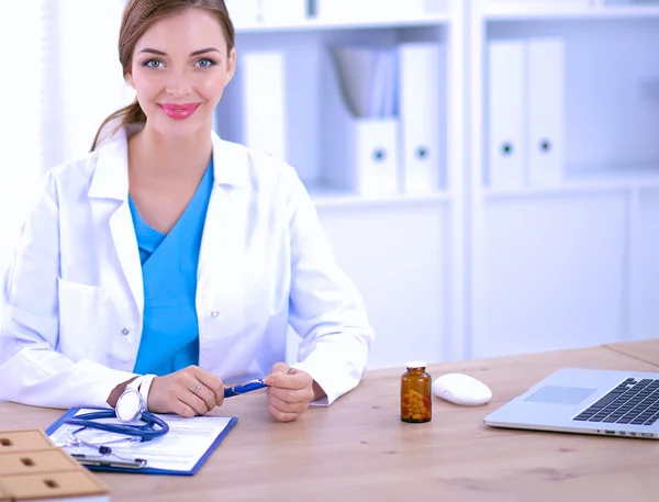 Beautiful young smiling female doctor sitting at the desk and writing.