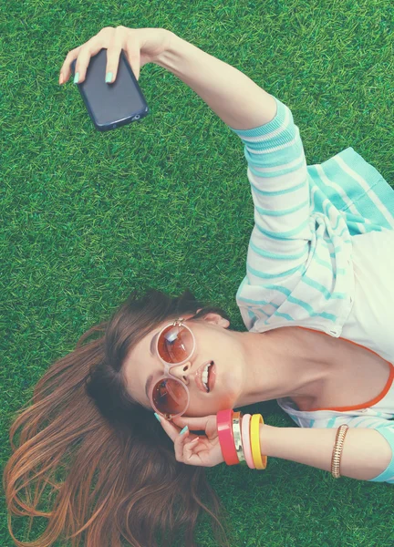 Beautiful young woman making selfie by her phone while lying in green grass