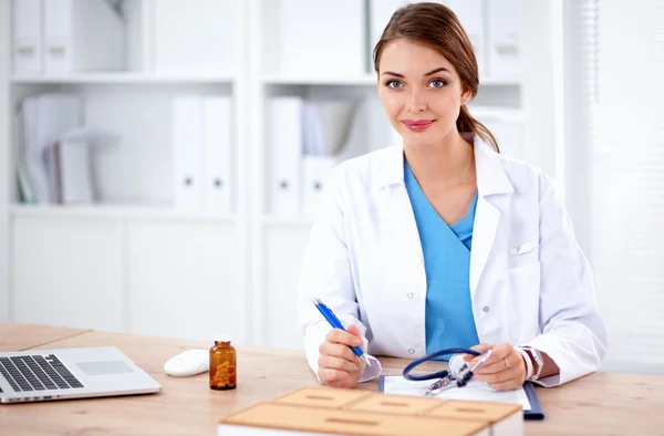 Beautiful young smiling female doctor sitting at the desk and writing.