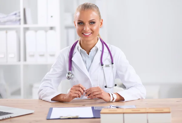 Beautiful young smiling female doctor sitting at the desk and writing.